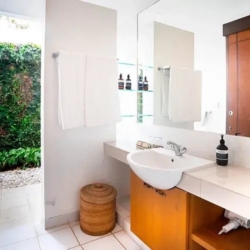 Asraya villa tropical guest bathroom with a clean white vanity large mirror and open doorway leading to a lush outdoor garden path sanur 870x500